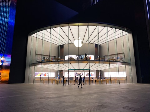 A modern, large glass-fronted store with an illuminated Apple logo on the facade. The interior features two levels visible through the clear glass with various Apple products on display. People are seen inside and outside the store, some walking and others standing.