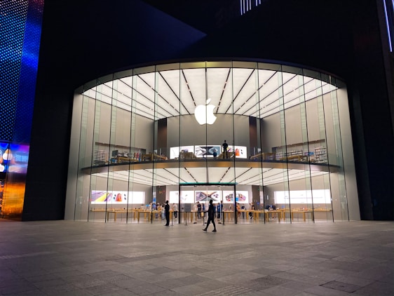 A modern, large glass-fronted store with an illuminated Apple logo on the facade. The interior features two levels visible through the clear glass with various Apple products on display. People are seen inside and outside the store, some walking and others standing.