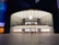 A modern, large glass-fronted store with an illuminated Apple logo on the facade. The interior features two levels visible through the clear glass with various Apple products on display. People are seen inside and outside the store, some walking and others standing.