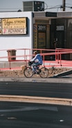 A person is riding a bicycle on a sidewalk next to a building with signs for auto sales and an insurance service. The building has a white exterior with red railings and several signs advertising services like DMV and taxes. The sidewalk is lined with rocks and small plants.