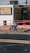 A person is riding a bicycle on a sidewalk next to a building with signs for auto sales and an insurance service. The building has a white exterior with red railings and several signs advertising services like DMV and taxes. The sidewalk is lined with rocks and small plants.