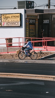 A person is riding a bicycle on a sidewalk next to a building with signs for auto sales and an insurance service. The building has a white exterior with red railings and several signs advertising services like DMV and taxes. The sidewalk is lined with rocks and small plants.