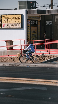 A person is riding a bicycle on a sidewalk next to a building with signs for auto sales and an insurance service. The building has a white exterior with red railings and several signs advertising services like DMV and taxes. The sidewalk is lined with rocks and small plants.