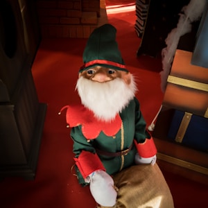 A festive elf figure is dressed in a green outfit with red accents and a fluffy white beard. It sits on a red floor, surrounded by holiday-themed decorations like brick walls and wooden structures. The scene is warmly lit, creating a cozy atmosphere.