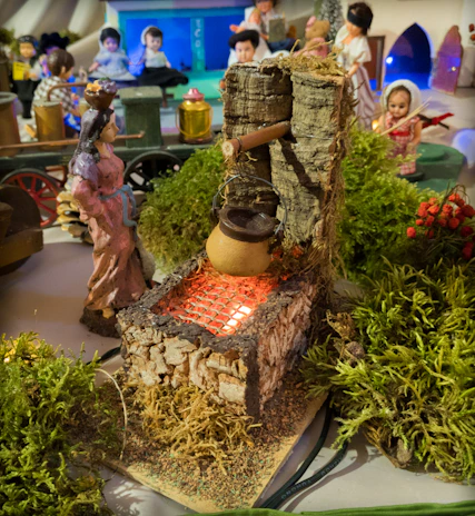 A miniature scene depicting a rustic setting with figurines and detailed elements. At the forefront, a small pot hangs over a simulated fire on a grill with red light mimicking heat, surrounded by green moss. Various figures are engaged in activities, with one prominently carrying a basket on her head. The background features a mix of more figurines and small architectural models.