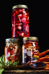Three glass jars filled with assorted pickled vegetables such as carrots, radishes, and possibly other root vegetables, arranged in a pyramid on a dark background. The jars are sealed with metallic lids, and the vegetables inside vary in color from deep red to orange and white. Fresh carrots with greens are placed next to the jars, and a wooden spoon is resting nearby.