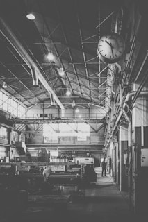 A large industrial workshop with high ceilings, exposed beams, and a variety of machinery. A clock is visible on the right side, indicating the time. There are overhead lights and large windows letting in natural light, with a few workers visible in the background.
