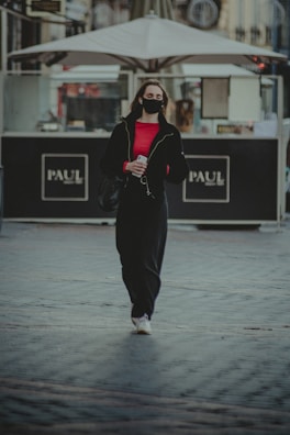 A person wearing a black mask and black clothing, with a bright red shirt underneath, is walking on a paved surface. They are holding a smartphone and a bag. Behind them, there is an outdoor seating area with an umbrella and signage that reads 'PAUL'. The background features a mix of urban architecture.