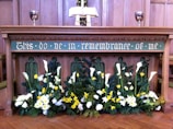 An altar with an inscription 'This do ye in remembrance of me' is decorated with an array of flowers, including white and yellow blooms, possibly lilies and daffodils. Two silver chalices are placed on either side of an open book on the altar, with wooden paneling in the background.