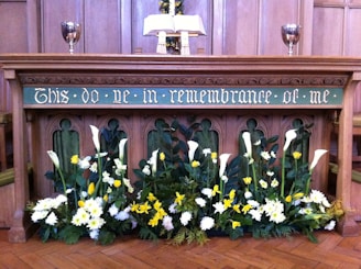 An altar with an inscription 'This do ye in remembrance of me' is decorated with an array of flowers, including white and yellow blooms, possibly lilies and daffodils. Two silver chalices are placed on either side of an open book on the altar, with wooden paneling in the background.