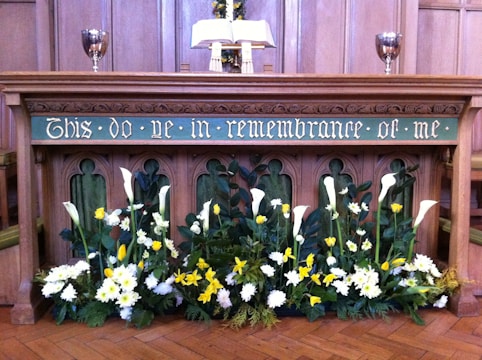 An altar with an inscription 'This do ye in remembrance of me' is decorated with an array of flowers, including white and yellow blooms, possibly lilies and daffodils. Two silver chalices are placed on either side of an open book on the altar, with wooden paneling in the background.
