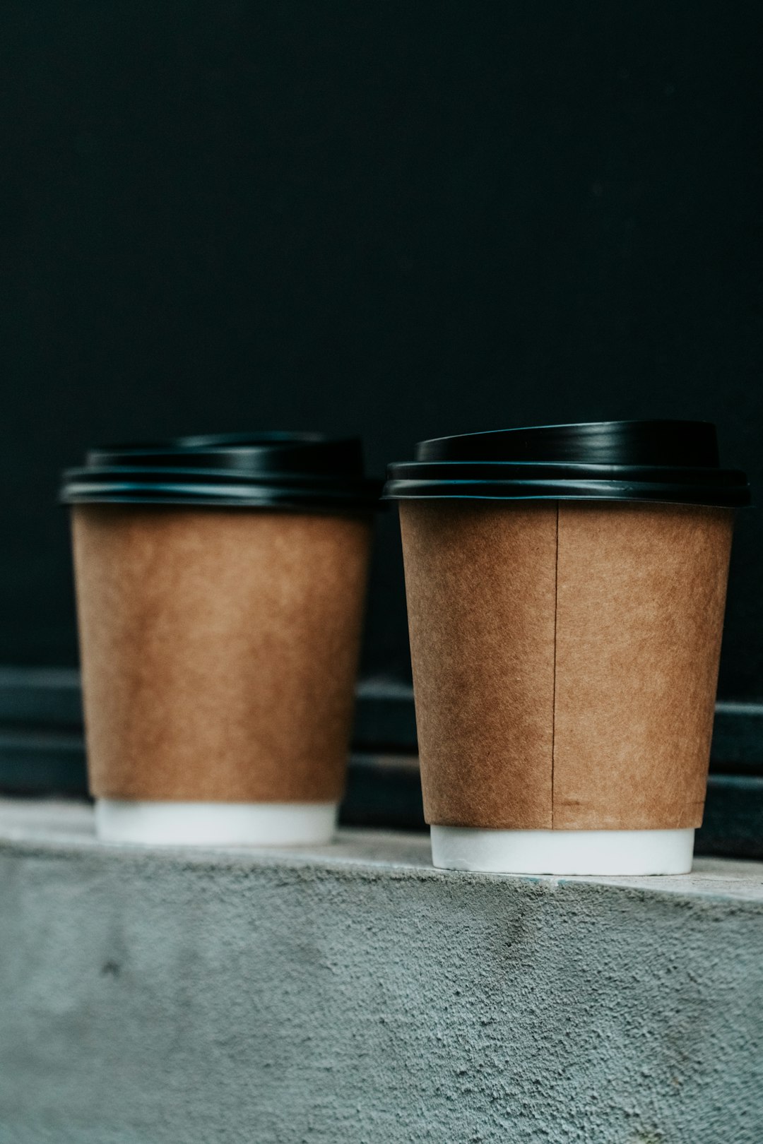 Two takeaway coffee cups with black lids on a concrete ledge