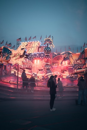 A vibrant carnival scene with bright neon lights and colorful rides set against a twilight sky. People are gathered around a spinning ride, and the atmosphere is lively and festive. Flags and signs add to the carnival ambiance.