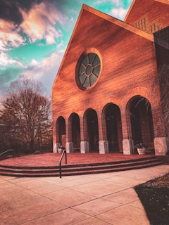 A large brick building features a prominent circular window and several tall arches. The architecture is traditional, with a slanted roof and detailed masonry work. The surrounding area includes bare trees and neatly laid out stone pathways, all under a vibrant sky with dynamic clouds.