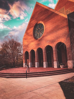 A large brick building features a prominent circular window and several tall arches. The architecture is traditional, with a slanted roof and detailed masonry work. The surrounding area includes bare trees and neatly laid out stone pathways, all under a vibrant sky with dynamic clouds.