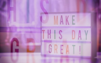 A motivational lightbox displaying the phrase 'MAKE THIS DAY GREAT!' with letters in pastel colors on a soft purple-tinted background. The image includes overlayed large letters adding a layered, dreamy effect.