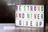 A lightbox with colorful letters displays the motivational message: 'Be Strong and Never Give Up'. The letters are in vibrant colors including green, magenta, and yellow, arranged across three lines. The background is softly blurred, giving a cozy and warm appearance.