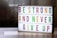 A lightbox with colorful letters displays the motivational message: 'Be Strong and Never Give Up'. The letters are in vibrant colors including green, magenta, and yellow, arranged across three lines. The background is softly blurred, giving a cozy and warm appearance.