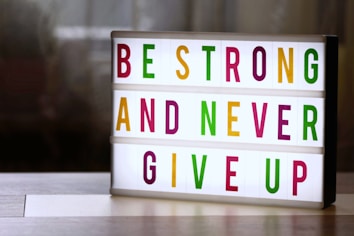 A lightbox with colorful letters displays the motivational message: 'Be Strong and Never Give Up'. The letters are in vibrant colors including green, magenta, and yellow, arranged across three lines. The background is softly blurred, giving a cozy and warm appearance.