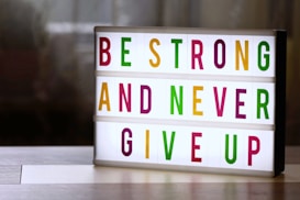 A lightbox with colorful letters displays the motivational message: 'Be Strong and Never Give Up'. The letters are in vibrant colors including green, magenta, and yellow, arranged across three lines. The background is softly blurred, giving a cozy and warm appearance.