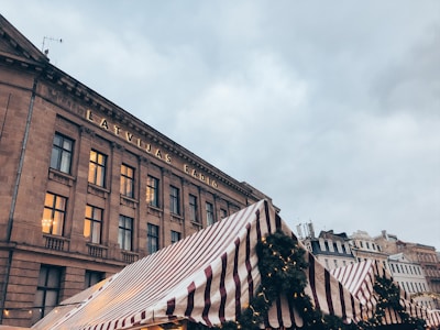 A historic building with the inscription 'Latvijas Radio' in large letters is prominently featured. The facade is illuminated with a warm glow from the windows. In the foreground, there is a striped canopy of a market stall, adorned with greenery and small lights. The surrounding sky is overcast, giving the scene a calm and subdued atmosphere.