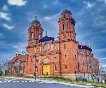 A large, historic red brick church with two prominent bell towers is situated on a corner with a clear road. The building features ornate architectural details and a grand entrance with a golden door. Flags are visible on a pole next to the church, and the sky above is filled with dramatic, dark clouds.