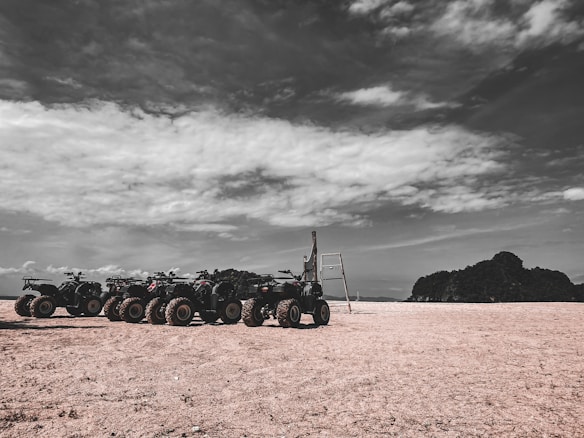 Several ATVs are parked in an open, sandy area under a partly cloudy sky. A rugged landscape with a solitary piece of wooden structure stands beside them. Hills can be seen in the distance.