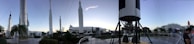 A panoramic view of a rocket garden displaying several space rockets, set in an outdoor museum environment. Tall, white rockets are stationed upright among palm trees and walkways, with a clear sky overhead. Visitors can be seen walking around, and the composition gives a sense of grandeur and human achievement in space exploration.