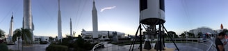 A panoramic view of a rocket garden displaying several space rockets, set in an outdoor museum environment. Tall, white rockets are stationed upright among palm trees and walkways, with a clear sky overhead. Visitors can be seen walking around, and the composition gives a sense of grandeur and human achievement in space exploration.