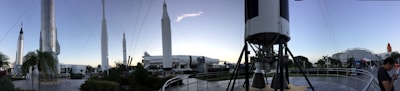 A panoramic view of a rocket garden displaying several space rockets, set in an outdoor museum environment. Tall, white rockets are stationed upright among palm trees and walkways, with a clear sky overhead. Visitors can be seen walking around, and the composition gives a sense of grandeur and human achievement in space exploration.