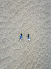 Two paddleboards with individuals on them are positioned side by side on calm, shallow water. The sandy texture of the sea bed is visible through the clear water, creating a tranquil and minimalistic scene.