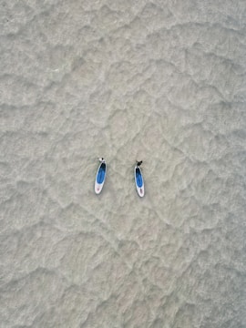 Two paddleboards with individuals on them are positioned side by side on calm, shallow water. The sandy texture of the sea bed is visible through the clear water, creating a tranquil and minimalistic scene.