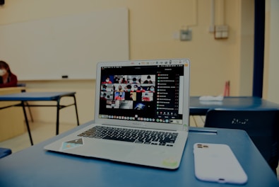 A classroom setting with a focus on a laptop on a desk, displaying a grid of video call participants. The room is sparsely furnished with blue chairs and a whiteboard visible in the background. A smartphone is placed beside the laptop.