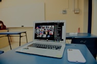 A classroom setting with a focus on a laptop on a desk, displaying a grid of video call participants. The room is sparsely furnished with blue chairs and a whiteboard visible in the background. A smartphone is placed beside the laptop.