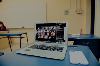 A classroom setting with a focus on a laptop on a desk, displaying a grid of video call participants. The room is sparsely furnished with blue chairs and a whiteboard visible in the background. A smartphone is placed beside the laptop.