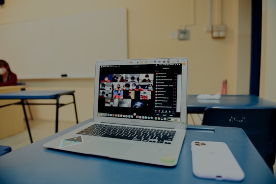 A classroom setting with a focus on a laptop on a desk, displaying a grid of video call participants. The room is sparsely furnished with blue chairs and a whiteboard visible in the background. A smartphone is placed beside the laptop.