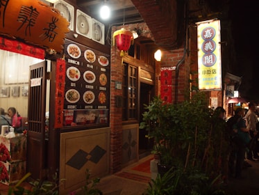A warmly lit street scene featuring a traditional Asian restaurant with brick walls and decorative lanterns. The entrance displays a menu with images of various dishes and signage in Chinese characters. People are visible near the entrance, adding to the lively atmosphere.