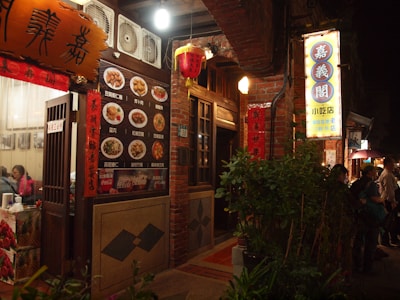 A warmly lit street scene featuring a traditional Asian restaurant with brick walls and decorative lanterns. The entrance displays a menu with images of various dishes and signage in Chinese characters. People are visible near the entrance, adding to the lively atmosphere.
