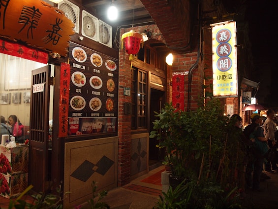 A warmly lit street scene featuring a traditional Asian restaurant with brick walls and decorative lanterns. The entrance displays a menu with images of various dishes and signage in Chinese characters. People are visible near the entrance, adding to the lively atmosphere.