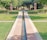 A landscaped garden with a long, narrow water channel leading towards two brown stone pillars. The surrounding area is lush with green grass and trimmed hedges, while trees provide a shaded backdrop. A pathway crosses the channel, flanked by neatly maintained bushes.