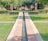 A landscaped garden with a long, narrow water channel leading towards two brown stone pillars. The surrounding area is lush with green grass and trimmed hedges, while trees provide a shaded backdrop. A pathway crosses the channel, flanked by neatly maintained bushes.