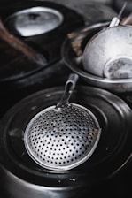A metallic strainer with numerous small holes rests inside a larger cooking pot. Other kitchen utensils and metal dishes are placed around, suggesting a kitchen setting. The materials are shiny and reflect light, indicating a well-used kitchen.