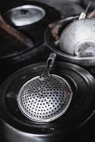 A metallic strainer with numerous small holes rests inside a larger cooking pot. Other kitchen utensils and metal dishes are placed around, suggesting a kitchen setting. The materials are shiny and reflect light, indicating a well-used kitchen.