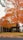 A residential house partially obscured by a large tree with vibrant orange autumn leaves. The tree's branches extend widely, with fallen leaves scattered across the lawn. An American flag hangs next to the house's entrance, and a wooden fence runs alongside the property.