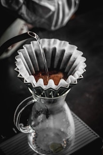 A pour-over coffee setup with a glass carafe and a filter holding coffee grounds. Hot water is being poured over the grounds from a kettle, and the setup is situated on a textured surface. The atmosphere is cozy and inviting, suggesting a quiet moment of coffee preparation.