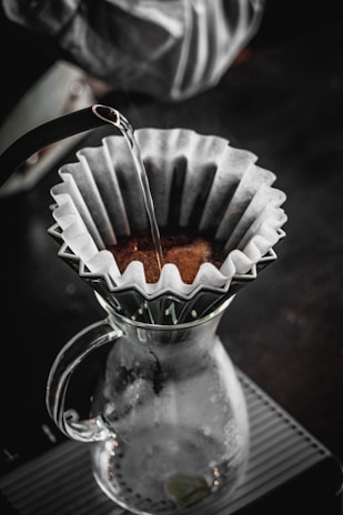 A pour-over coffee setup with a glass carafe and a filter holding coffee grounds. Hot water is being poured over the grounds from a kettle, and the setup is situated on a textured surface. The atmosphere is cozy and inviting, suggesting a quiet moment of coffee preparation.