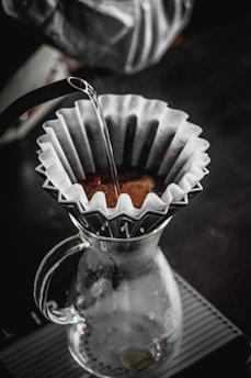 A pour-over coffee setup with a glass carafe and a filter holding coffee grounds. Hot water is being poured over the grounds from a kettle, and the setup is situated on a textured surface. The atmosphere is cozy and inviting, suggesting a quiet moment of coffee preparation.