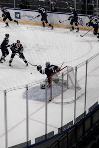 Several ice hockey players are skating and practicing on a rink. A goalie is positioned in front of the net, defending against incoming pucks. The scene is surrounded by boards and transparent protective glass, with stadium seating visible in the background.