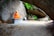 A person dressed in bright orange robes sits cross-legged in meditation on a stone bench against a large rock wall. The scene is set outdoors, beneath an arch formed by a massive boulder, with greenery visible in the background. There are some signs nearby on the ground.