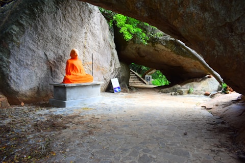 A person dressed in bright orange robes sits cross-legged in meditation on a stone bench against a large rock wall. The scene is set outdoors, beneath an arch formed by a massive boulder, with greenery visible in the background. There are some signs nearby on the ground.